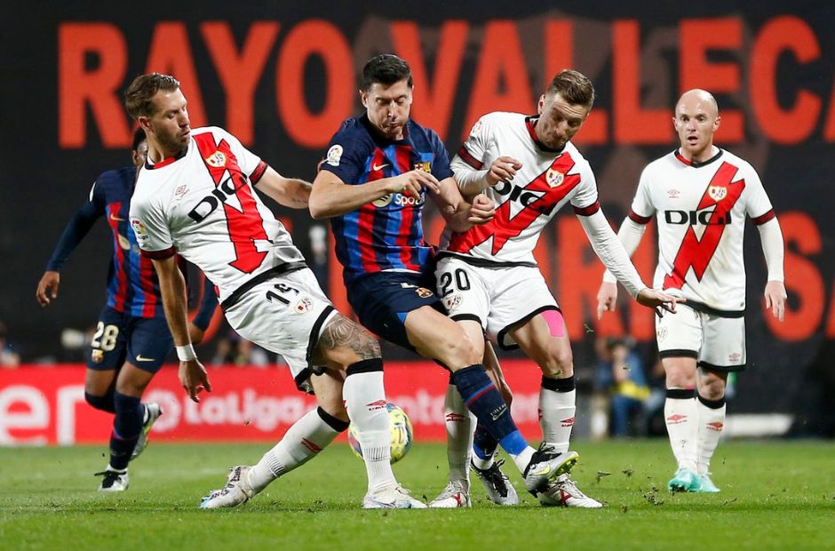Các cầu thủ Rayo trong trang phục quần áo trắng, sọc chéo đỏ đang tranh chấp bóng cùng tiền đạo Lewandowski của Barca.